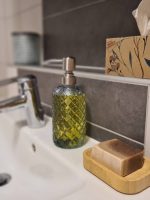 Eine feste Stückseife auf einer Holzablage und ein grüner Glas-Seifenspender neben einer modernen Armatur am weißen Waschbecken.