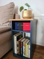 Ein blauer Beistelltisch neben einem beigen Sofa, bestückt mit verschiedenen Büchern, drei Boxen des Fragespiels Sondermoment und einer kleinen Pflanze in einem goldenen Topf.