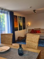 Blick über einen Holzesstisch mit blauen Vasen auf ein gemütliches Sofa, einen blauen Teppich und ein Regal voller Brettspiele.