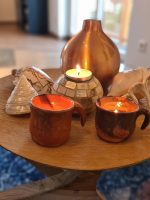 Nahaufnahme eines runden Holztabletts auf einem Glastisch mit zwei handgetöpferten Tassen, brennenden Kerzen, dekorativen Muscheln und einer kupferfarbenen Vase.