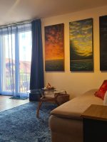 Gemütliches graubraunes Sofa mit roten Kissen, davor ein runder Glastisch mit brennender Kerze und drei handgemalte Ölgemälde an der Wand.