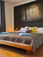 Ein großes Doppelbett mit grauer Tagesdecke, bunten Zierkissen und frisch gefalteten blauen Handtüchern vor einer dunkelgrauen Wand mit einem großen Waldbild.