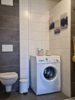 Gesamtansicht eines modernen Badezimmers mit Hänge-WC, Waschmaschine, Deko-Körben, einer verglasten Dusche und dekorativen Waldbildern an den hellen Fliesen.