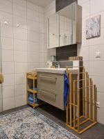 Modernes Badezimmer mit einem beigen Waschtisch, großem dreitürigen Spiegelschrank, Handtuchhaltern aus Holz und einem orientalisch gemusterten Teppich.