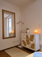 Elegante weiße Kommode mit goldenen Blattmotiven, einer leuchtenden Salzlampe und einem großen Spiegel mit prunkvollem Goldrahmen im hellen Schlafzimmer der Ferienwohnung.