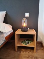 Eine eingeschaltete Design-Lampe mit warmem Licht steht auf einem hölzernen Nachttisch vor einer dunkelgrauen Wand neben dem Bett.