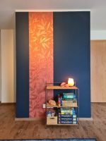 Gesamtansicht einer Designwand mit Spiele-Ecke mit einem Rattanregal vor einer dunkelblauen Wand mit einer rot-gold gemusterten Tapetenbahn im Hintergrund.