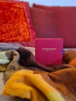 Das rote Kartenspiel Sondermoment Love Edition liegt liebevoll drapiert in einer weichen Wolldecke vor roten Sofakissen.