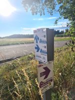 Ein offizieller weiß-lila Wanderwegweiser des Innviertels für den Adelsberg Weg und den Höhwirt Weg an einer sonnenbeschienenen Wiese.