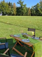 Detailansicht des festlich gedeckten Tisches mit grünem Tischläufer und Geschirr auf der sonnigen Wiese im Garten Perle. Gedeckter Gartentisch mit Tischläufer und Dekoration im Garten Perle am Kobernaußerwald