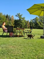 Frau sitzt an einem gedeckten Gartentisch mit grünem Sonnenschirm im weitläufigen Garten am Waldrand