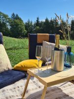 Gemütliches Picknick-Set mit Kissen, Holztischchen und Sektkühler auf einer Decke im Gras